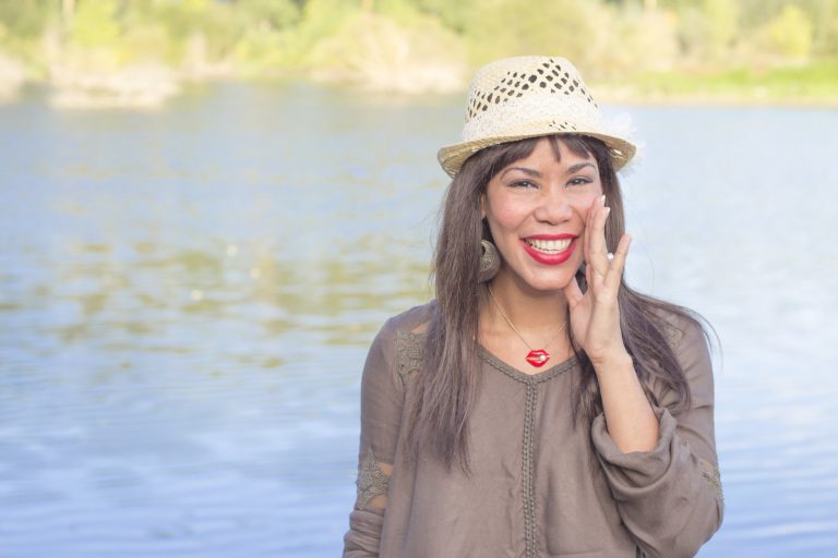 Smiling young woman with an open hand to the side of the mouth and a lake in the background.